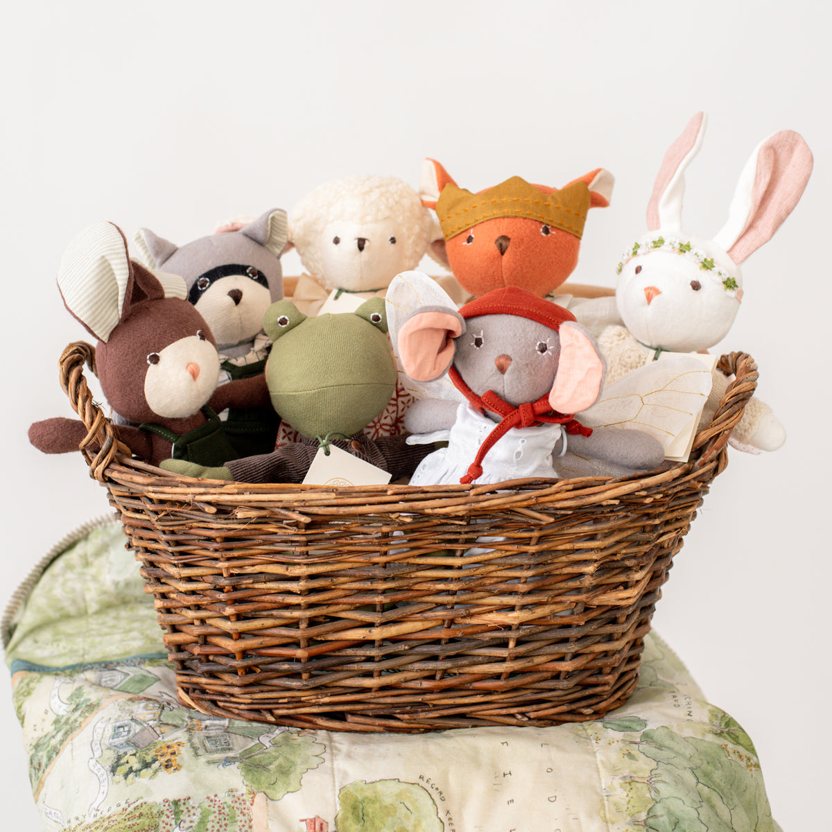 A wicker basket filled with Hazel Village animals who are wearing charming outfits. The basket rests atop a blanket which is printed with watercolor artwork of the Hazel Village map. 